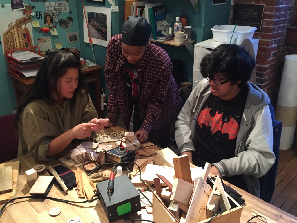 a teaching artist shows two teens how to use woodburning tools